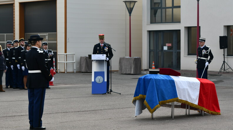 Au premier plan, une gendarme en tenue de c&eacute;r&eacute;monie, vue de trois-quarts dos. Devant elle, &agrave; droite de l'image, un cercueil pos&eacute; en hauteur, rev&ecirc;tu du drapeau tricolore, sur lequel sont pos&eacute;s un k&eacute;pi et un coussin rouge. En arri&egrave;re plan, &agrave; gauche, des gendarmes au garde-&agrave;-vous ; au centre, un g&eacute;n&eacute;ral derri&egrave;re un pupitre, prononce une allocution au micro ; &agrave; gauche, un gendarme seul au garde-&agrave;-vous, sabre &agrave; l'&eacute;paule.