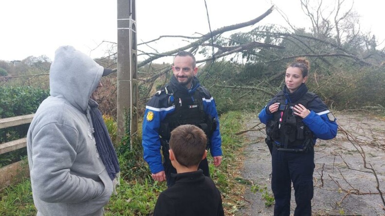 Un homme et une femme gendarmes, en tenue, debout, de face, sur la photo, disucutent avec un homme que l'on voit de biais, emitouflé dans un sweat à capucje gris, avec à ses côtés, un jeune garçon, qui se tient dos au photographe. Derrière eux, on aperçoit le ciel gros et des arbres couchés après la tempête, ainsi qu'un poteau électrique