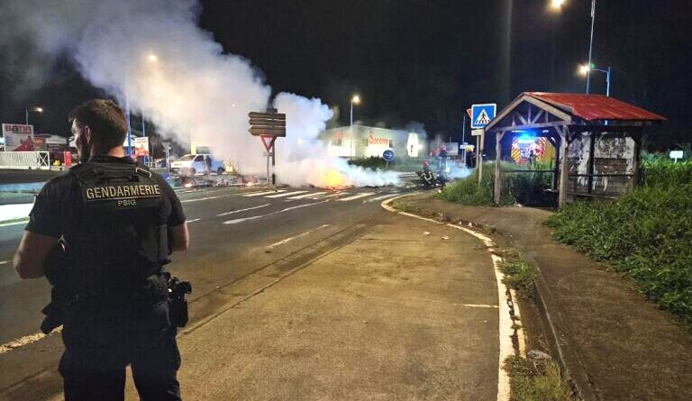 Un gendarme se tient debout, de dos, en uniforme, dans la rue, de nuit. ll fait face à un incendie déclenché sur la chaussée, dont s'échappe une grosse fumée blanche.