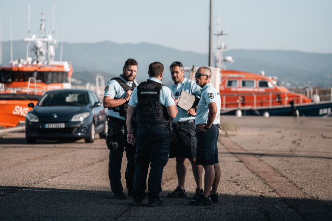 Agrandir l'image Des gendarmes discutent dans un port. On aperçoit en arrière plan deux bateaux de couleur orange.