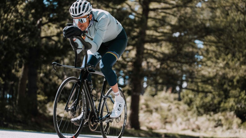 Un cycliste avance sur son v&eacute;lo, le corps pench&eacute; en avant, gravissant une c&ocirc;te. V&ecirc;tu d'une tenue bleue, il porte un casque, des lunettes de soleil blanches et des gants noirs.