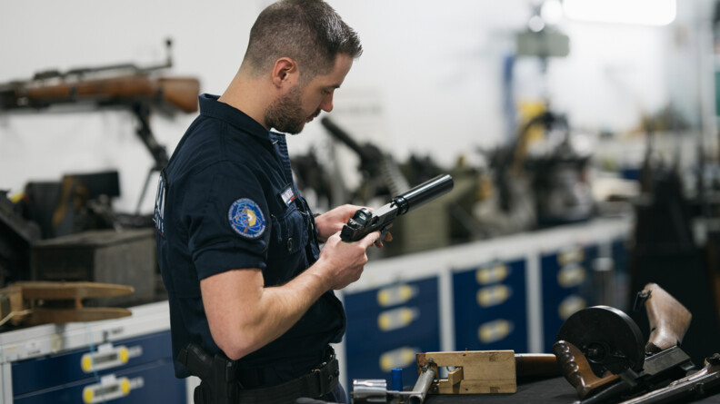 Dans une pièce éclairée à la lumière artificielle, un homme se tient debout, de profil, devant une table noire. Sur celle-ci sont posées plusiers armes. L'homme, vêtu d'un uniforme bleu marine, et portant une arme à la ceinture; manipule une arme, qu'il tient des deux mains. A l'arrière, on aperçoit un grand meuble bleu marine à tiroirs qui fait toute la largeur de la photo, sur lequel sont posées des boîtes et des armes.