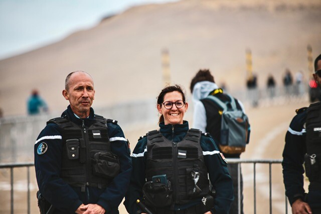 Deux gendarmes de face, souriants, et un troisième à droite, dont on ne voit que la moitié, sont debout devant une barrière. Derrière eux on devine des gens à pied et le paysage minéral du Mont Ventoux.