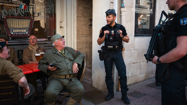 Deux gendarmes échangent dans la rue avec des hommes, habillés en tenue militaire pour l'occasion et assis en terrasse.