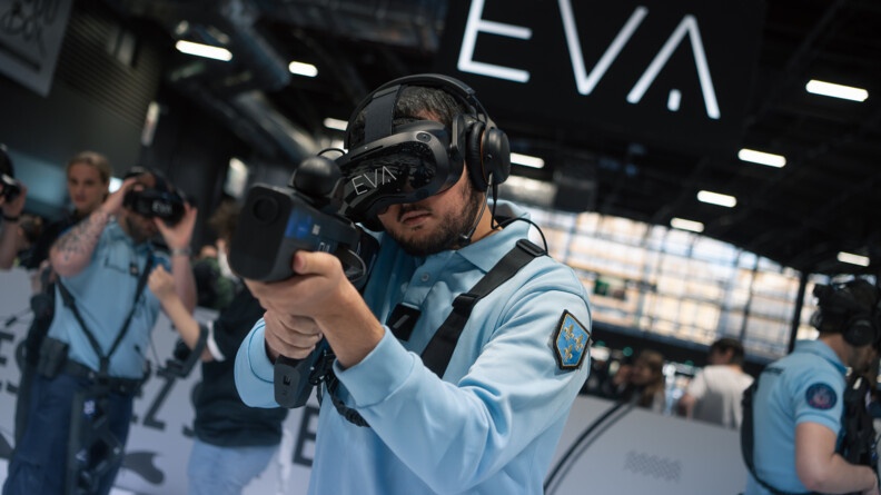 Dans un grand hall, un gendarme en tenue tient une arme devant lui, avec ses deux mains, à hauteur d'épaules. Il porte un casque de réalité virtuelle devant les yeux.