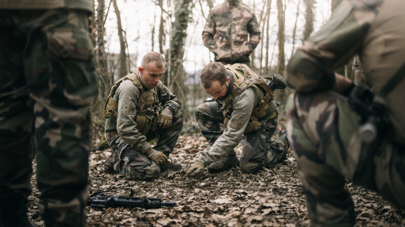 Deux militaires en treillis au sol expliquent une manoeuvre. Quatre militaires en treillis debout les observent