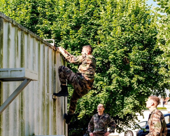 Agrandir l'image Un élève militaire en tenue camouflage escalade un mur en taule, devant d'autres militaires positionnés au pied du mur.