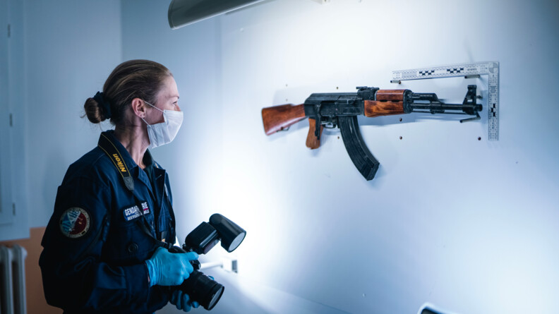 Une femme gendarme, équipée de gants bleus, d'un masque et tenant un appareil photo dans les mains, se tient face à un fusil, supendu sur un mur blanc, et éclairé par une lampe murale située au dessus