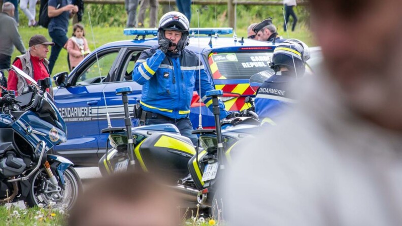 Deux personnages flous au premier plan. Au second plan, deux motocyclistes d'EDSR, l'un de face, debout à côté de sa moto, en train de parler dans son micro ; le second, vu de dos, sur sa moto. En arrière plan, une moto et une voiture gendarmerie et du public.