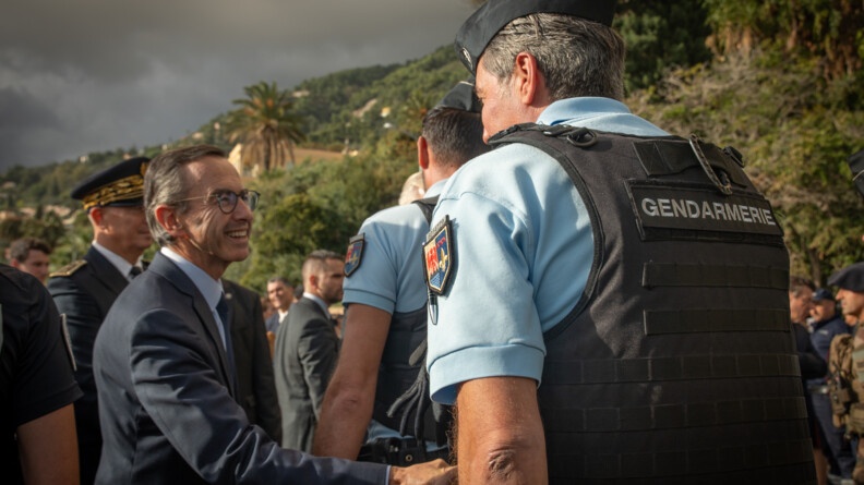 Le ministre de l'Int&eacute;rieur, Bruno Retailleau, serre la main &agrave; un gendarme, que l'on voit de dos. Ce dernier porte un polo bleu ciel et un gilet par balle noir, portant dans le dos l'inscription "Gendarmerie"