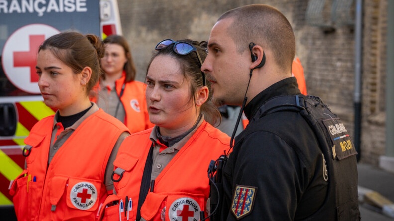 Photo d'un gendarme, aux c&ocirc;t&eacute;s de deux profesionnelles de la croix rouge fran&ccedil;aise. On aper&ccedil;oit le v&eacute;hicule de la Croix rouge en arri&egrave;re plan.