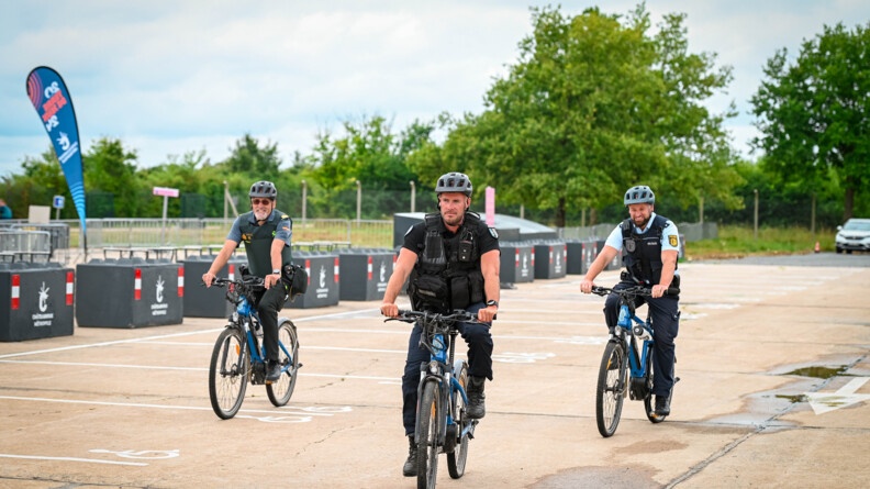 Un gendarme français au centre, avec, au second plan, à gauche, un garde civil espagnol, et à droite, un policier allemand, tous deux souriants, patrouillent à vélo en formation "en pointe",  sur parking désert.