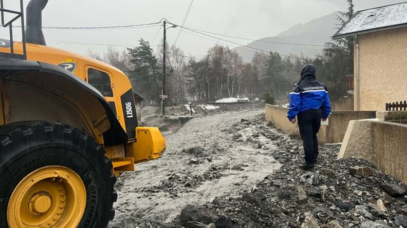 Au premier plan, à gauche, l'avant d'un engin de chantier jaune. Au second plan, à droite, un gendarme de dos, vêtu d'un blouson de pluie, capuche sur la tête, sur une digue de fortune érigée à proximité d'un fleuve de boue dévalant au milieu des habitations.