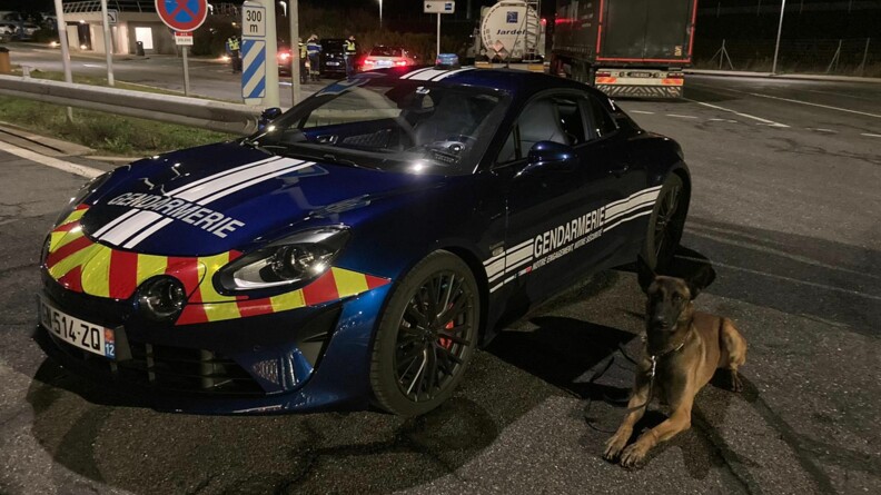 A gauche, un véhicule rapide d'intervention (VRI) Alpine 110 de la gendarmerie, à droite, sagement couché, un chien d'une équipe cynophile. En arrière plan, les gendarmes contrôlent des véhicules et des poids lourds.