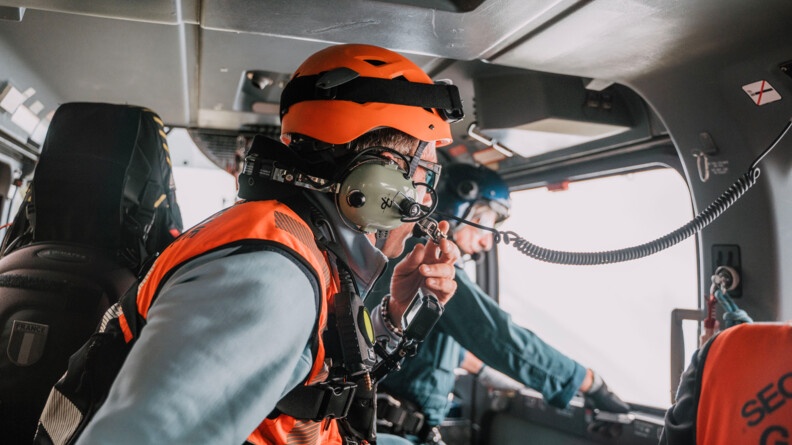Vue de l'intérieur d'un cockpit d'hélicoptère gendarmerie, deux gendarmes sont un vếtu d'une chasuble orange. Au fond l'autre, flou, en combinaison de vol, s'apprête à ouvrir la porte latérale