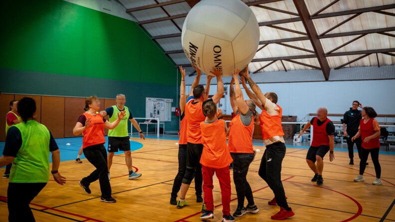 Vue de dos sur un groupe d'hommes et de femmes de tous âges, portant une chasuble orange ou jaune fluo, jouent sur le parquet d'un gymnase. Au centre, cinq d'entre eux essaient de lancer un gros ballon de volley gonflable.