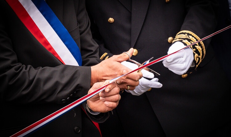 Photo représentant les mains de deux personnes, dont l'une porte des gants blancs, et coupent ensemble un ruban tricolore aux couleurs de la France. l'une des personnes porte un uniforme noir, dont le bout des manches sont gansées de broderies dorées, tandis que la seconde (un élu), porte un costume noir et une écharpe tricolore sur la poitrine.