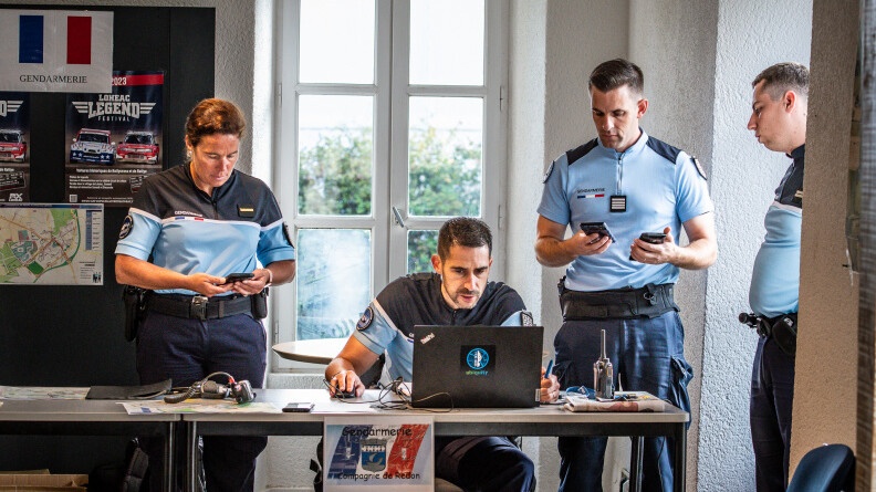 Derrière une table, dans une salle avec une fenêtre, 4 gendarmes, trois hommes et une femmme à gauche. Un des hommes est assis derrire un ordinateur portable posé sur la table. Les deux autres gendarmes sont à droite et regardent vers l'écran