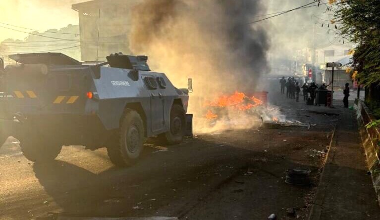 Un véhicule blindé de la gendarmerie, de couleur bleue, circule sur une route, se dirigeant droit sur une barricade en feu barrant la voie, dont s'échappe une épaisse fumée noire montant en dircetion du ciel.