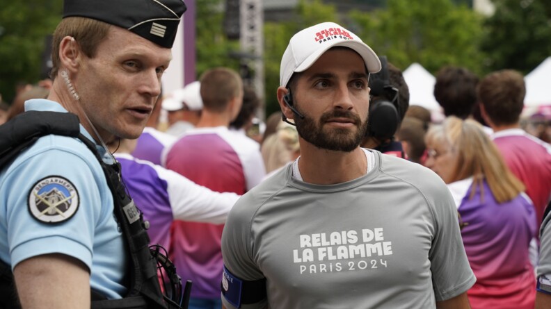 Agrandir l'image Un gauche un chef d'escadron de gendarmerie et à droite un runner arborant un t-shirt gris "relais de la flamme paris 2024" et un brassard au bras droit de la gendarmerie. Derrière eux, la foule