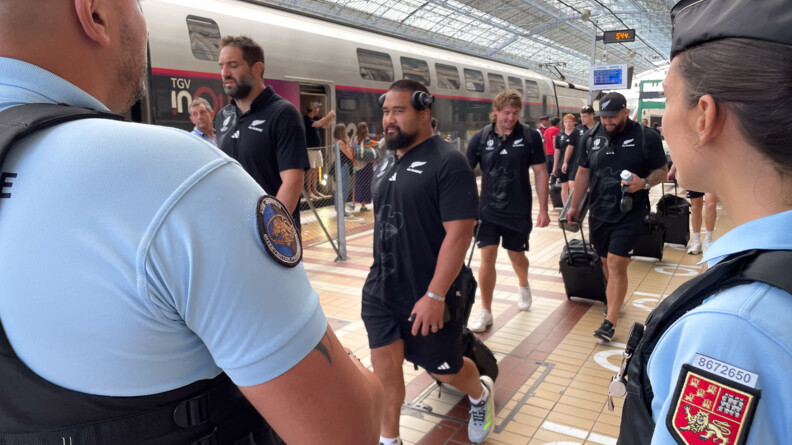 Au premier plan, un gendarme à gauche, vu de trois quart dos, et une gendarme, à droite, vue de profil, surveillent l'arrivée des All Blacks sur le quai de la gare. Au second plan, des joueurs de l'équipe néozélandaise, en short et tee-shirt noirs,  transitent avec leurs bagages. En arrière plan, du public les observe derrière une paroi vitrée et en fond un TGV.