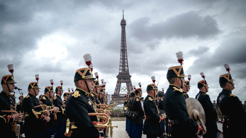 photo de la Tour Eiffel revêtant les anneaux olympiques. Devant elle, de biais, se tiennent des gardes républicains.