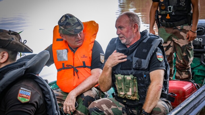 Le DGGN en polo noir pantalon de treillis chapeau de brousse sur la tête et portant un gilet de sauvetage orange est assis dans une pirogue. Il discute avec un gendarme assis à côté de lui. Derrière eux on voit un gendarme debout et devant eux on voit l'épaule d'un gendarme assis.