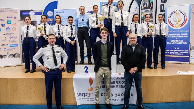 Agrandir l'image 11 coureuses en tenue chemise blanche et pantalon bleu avec leurs médailles sur la scène des prix et le podium. En fond des kakémonos des sponsors et de la gendarmerie. Au premier plan au pied de la scène, à gauche un général deux étoiles de gendarmerie dans la même tenue que les coureuses et deux hommes en civil