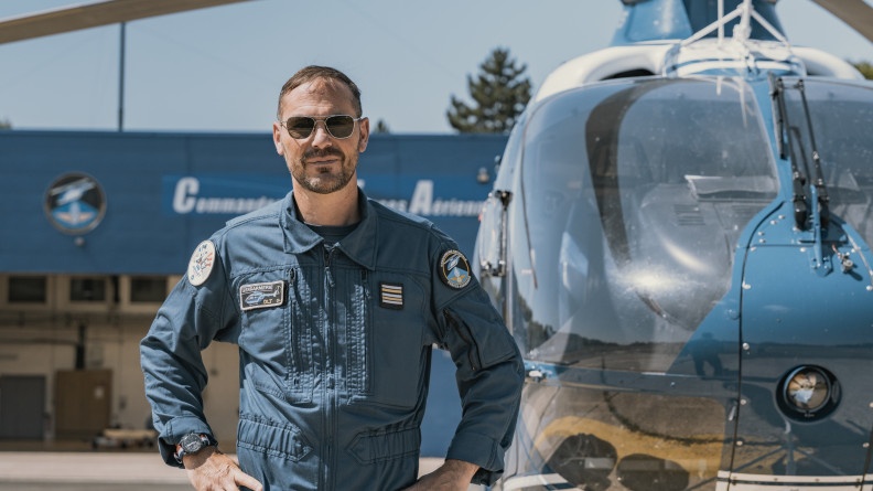 Agrandir l'image La photo représente un pilote d'hélicoptère, posant debout, mains sur les hanches, devant l'hélicoptère bleu de la gendarmerie.