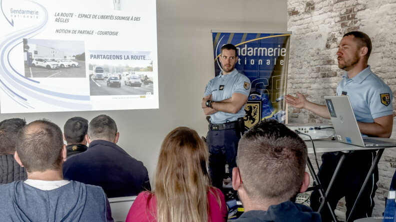 Poursuite de la présentation par deux gendarmes debout dans une salle, sur la droite, aux personnes assises sur des chaises. Le power point projeté indique "partageons la route"