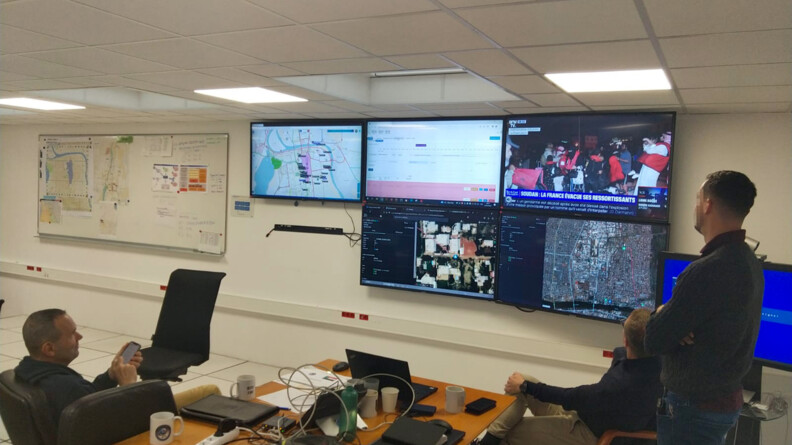 Salle de crise du GIGN montrant plusieurs &eacute;crans sur le mur du fond et un grand tableau blanc &agrave; gauche . Des ordinateurs et des t&eacute;l&eacute;phones sont pos&eacute;s sur une table au centre. A gauche, le commandant du GIGN assis, t&eacute;l&eacute;phone en main, regarde les &eacute;crans. A droite, un autre personnel assis et un debout les regardent &eacute;galement.