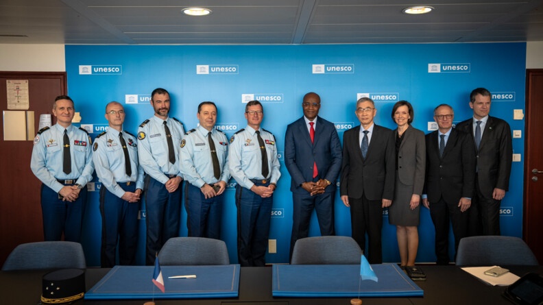 Agrandir l'image A gauche 5 gendarmes et à droite 5 civils dont une femme au milieu, derrière une table noire et devant un mur bleu siglé UNESCO