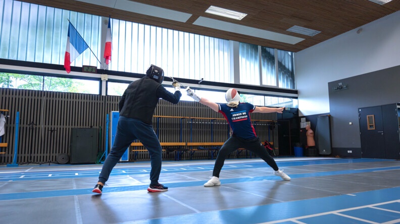 Vue de profil de deux escrimeurs, à gauche il est vêtu de noir et réalise une parade face à son adversaire, vêtu de bleu et rouge, qui s'étend pour le toucher