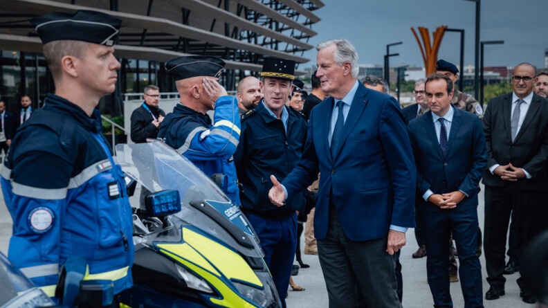 Le premier ministre, Michel Barnier, tend la main vers un gendarme en faction devant le stade de France (Saint-Denis). D'autres personnalités officielles se tiennent à leurs côtés.