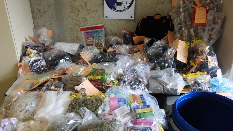 De nombreux produits stupéfiants, disposés dans des sacs plastique, sont rassemblés sur une table. Sur le mur derrière, une pancarte indique "Gendarmerie nationale - Compagnie de Brest"