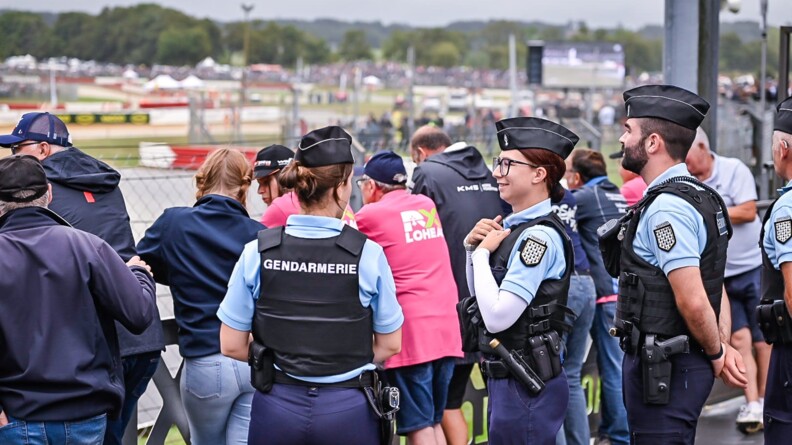 Une gendarme de dos et deux de profil, sur la droite de la première. Derrière eux, le public accoudé aux barrières de course. EN arrière-plan, le village de course, flou