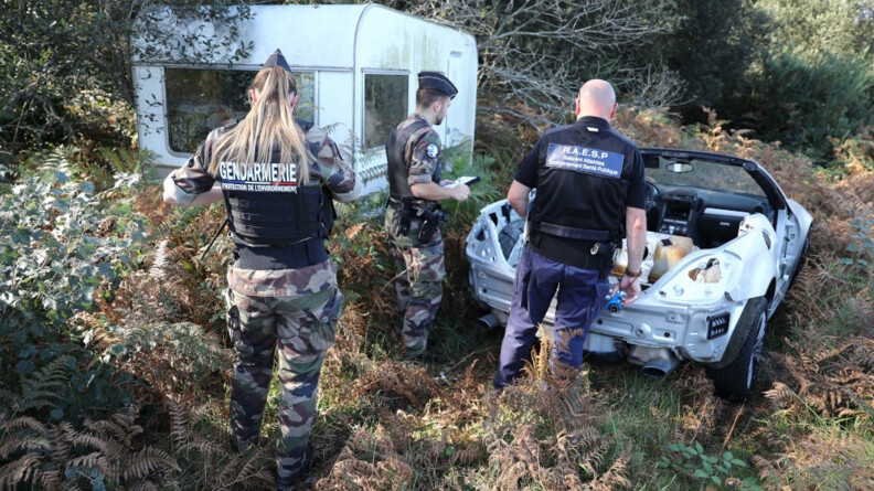 Agrandir l'image Deux gendarmes en treillis, porteurs d'un gilet pare-balle, et un troisième militaire, référent atteintes à l'environnement et à la santé publique, vus de dos, devant l'épave d'un cabriolet blanc partiellement désossé, déposé au milieu des fougères, à l'orée d'un bois, juste à côté d'une vieille caravane blanche.