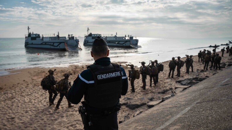 Agrandir l'image Un gendarme de dos regarde la plage où vient de se dérouler la reconstitution du débarquement. En fond, on observe deux bateaux ainsi que des militaires en tenue.