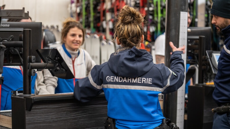 Au premier plan, une femme gendarme se tient debout, de dos. Elle a les cheveux châtains, relevés en chignon, et parle en levant la main droite, regardant la femme - une commerçante - se tenant face à elle, le regard tourné vers l'objectif. Elle sourit. A sa droite, on aperçoit du matériel informatique. Et derrière elle, du matériel de ski.