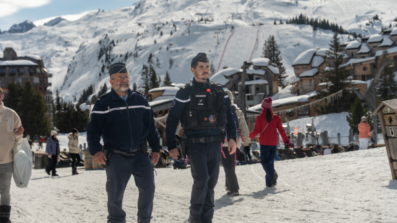 Agrandir l'image Deux gendarmes marchent côte à côte dans une rue enneigée d'une station de ski, au milieu de touristes. Derrière eux se dresse la montagne sur laquelle on aperçoit les remontées mécaniques. Le temps est ensolleilé.