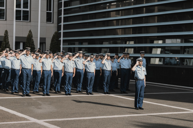 Des gendarmes, align&eacute;s c&ocirc;te &agrave; c&ocirc;te, et les uns derri&egrave;re les autres, effectuent le salut militaire. La sc&egrave;ne se d&eacute;roule de jour.