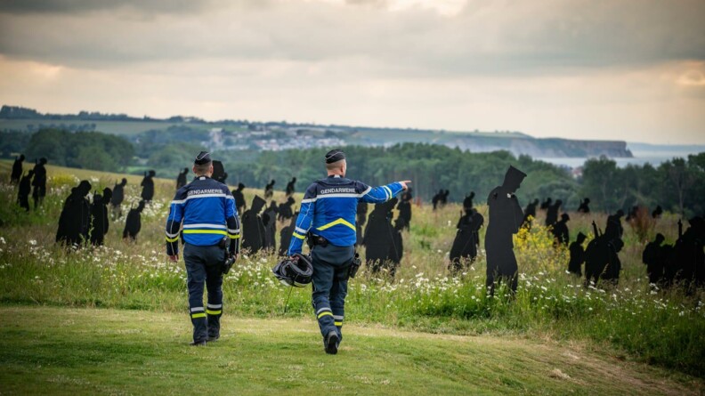 Deux gendarmes marchent sur une pelouse et en fond se trouve des silhouettes noirs.