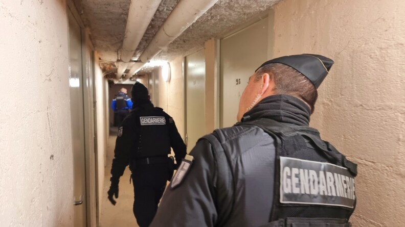 Deux gendarmes mobiles et un gendarme départemental dans un couloir blanc des parties communes au sous-sol d'un immeuble