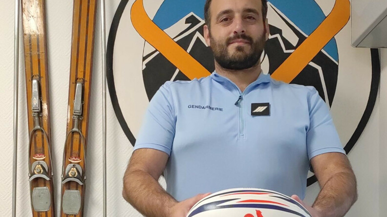 Portrait en buste d'un gendarme barbu présentant dans ses mains un ballon de France rugby. A droite, des skis anciens accrochés au mur. Derrière lui, une rondache taille humaine de la gendarmerie de montagne