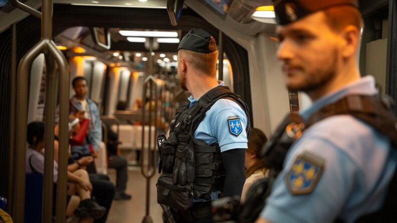 Au premier plan, un gendarme de profil, flou, regarde droit devant lui. Derrière lui, un second gendarme, également de profil, a la tête tournée vers l'arrière de la voiture de métro, où l'on voit des personnes assises en arrière-plan.