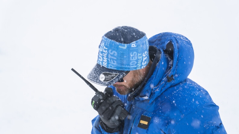 Agrandir l'image Dans un paysage enneigé, un homme vếtu d'un anorak et d'une casquette de couleur bleue, dont une aperçoit uniquement le haut du corps, se tient debout, la tête inclinée vers l'avant. Il tient dans sa main droite un talkie walkie