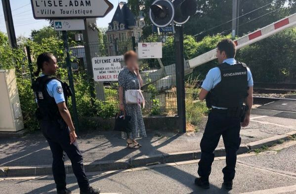 Un gendarme vu de profil et un gendarme vu de dos dispensent des conseils de pr&eacute;vention &agrave; une passante, &agrave; proximit&eacute; d'un passage &agrave; niveau, dont les barri&egrave;res sont en train de descendre.