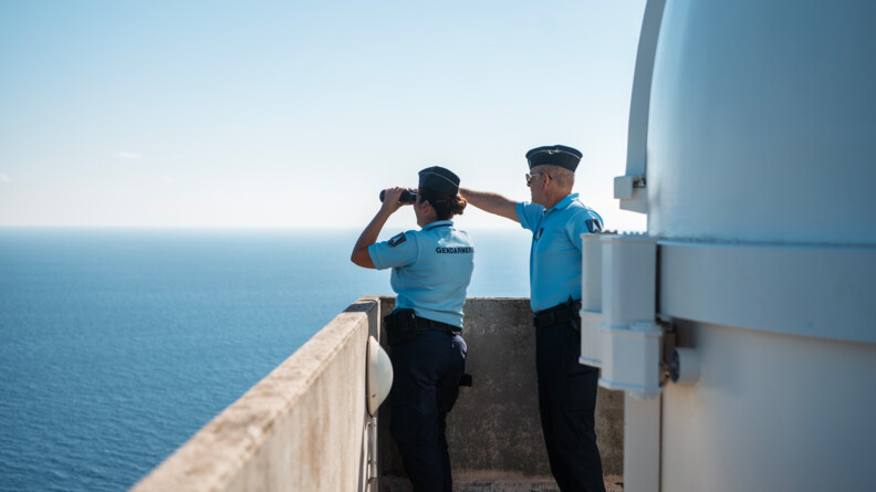 Sur un point de vue en hauteur, deux militaires observent le large. L'une regarde avec des jumelles alors que son camarade lui montre quelque chose au loin.