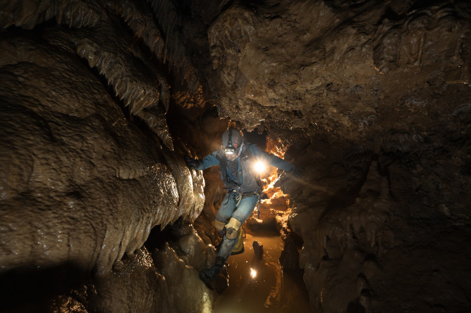 Agrandir l'image Photo montrant un gendarme spéléologue équipé d’une combinaison, d’un casque et d’une lampe frontale, progressant prudemment dans un passage étroit et humide d’une grotte. équilibre, les mains appuyées sur les parois rocheuses, au-dessus d’un petit plan d’eau reflétant la lumière.