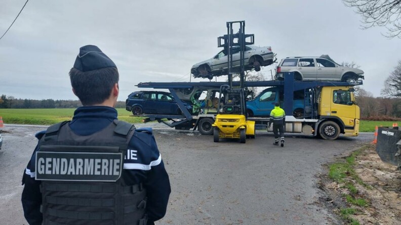 Au premier plan, un gendarme de dos. Au second plan, un chariot élévateur jaune charge des épaves de véhicules sur un camion à tête jaune.